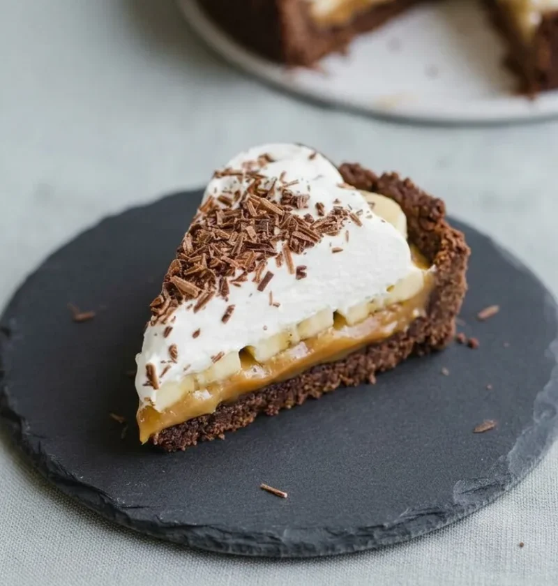 Slice of banoffee pie showing chocolate Hobnob base, toffee, banana, and cream layers with chocolate shavings