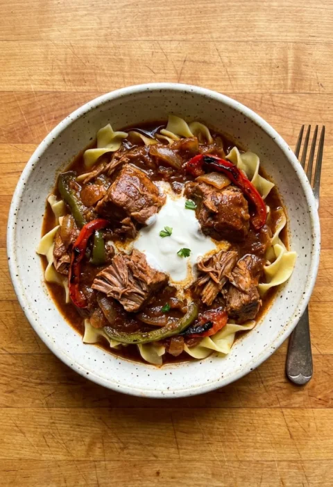 Pork goulash with falling-apart shoulder peppers and soured cream over egg noodles in a speckled bowl