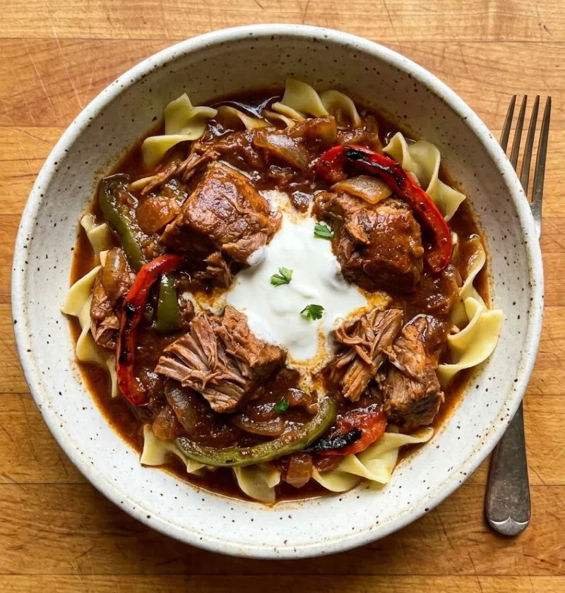 Pork goulash with falling-apart shoulder peppers and soured cream over egg noodles in a speckled bowl