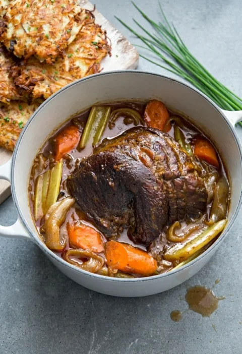 Hairy Bikers beef brisket pot roast, whole browned joint in a casserole with carrots, celery and onion wedges in dark gravy, potato latkes behind