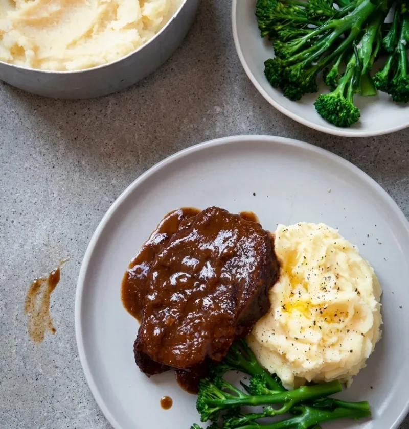 Hairy Bikers braised beef, a glazed piece of feather blade in glossy red wine sauce with buttery mashed potato and tenderstem broccoli on a grey plate