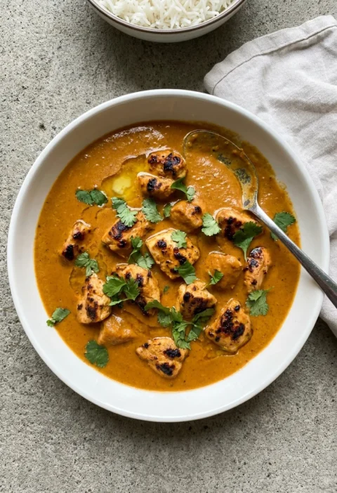 Butter chicken makhani in a white bowl with grilled chicken pieces in a smooth amber-orange ghee and cream sauce topped with fresh coriander and a bowl of rice