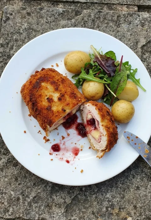 Chicken cordon bleu cut in half on a white plate showing layers of ham, melted Emmental, and cranberry sauce inside a golden double-breaded crust with new potatoes and salad