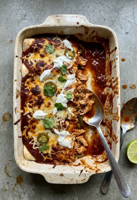 Chicken enchiladas in a baking dish with dark red chipotle sauce, melted Cheddar, soured cream, coriander, and jalapeños with one portion scooped out showing the filling