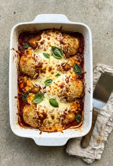 Chicken parmo parmigiana in a white baking dish with breaded chicken under melted browned mozzarella on a red tomato sauce with fresh basil leaves