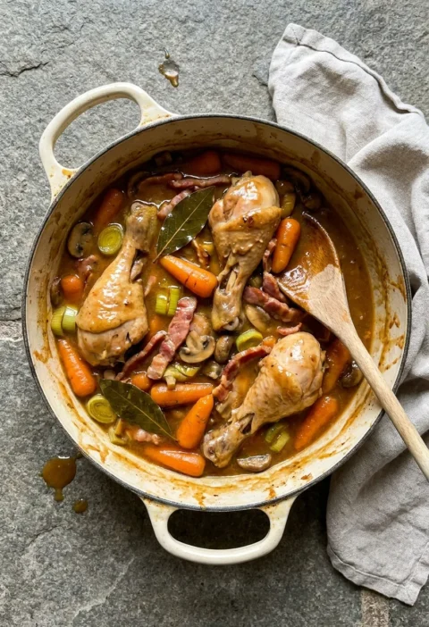 Old-fashioned chicken stew in a casserole dish with bone-in chicken pieces, Chantenay carrots, leek slices, mushrooms, and bacon in a thick cider sauce