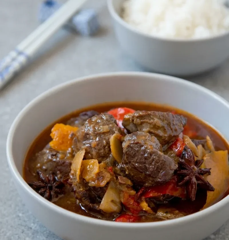 Hairy Bikers Chinese beef curry, tender chunks of beef shin with red peppers in a rich reddish-brown sauce with whole star anise and orange peel in a grey bowl, rice behind
