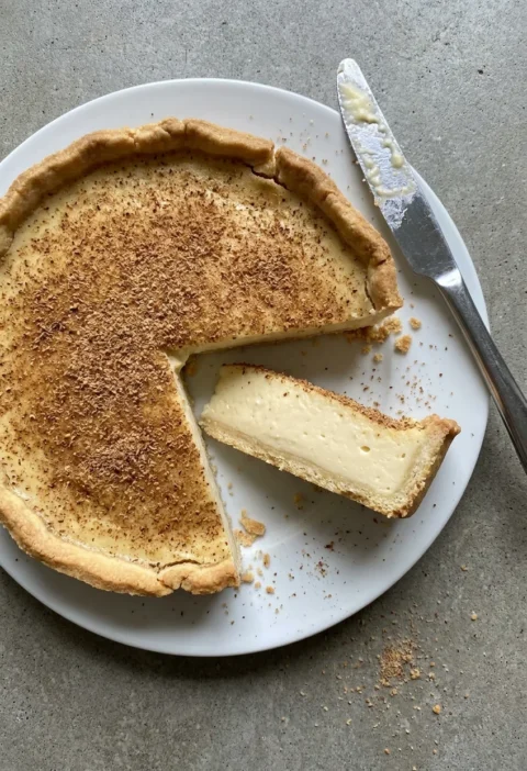 Hairy Bikers custard tart with a nutmeg covered top in golden shortcrust pastry, one slice cut showing the thick set custard