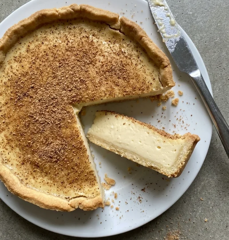 Hairy Bikers custard tart with a nutmeg covered top in golden shortcrust pastry, one slice cut showing the thick set custard