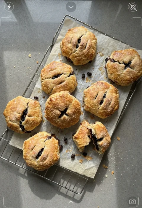 Hairy Bikers eccles cakes cooling on a wire rack with sugar glazed tops, knife slashes showing the currant filling and one broken open