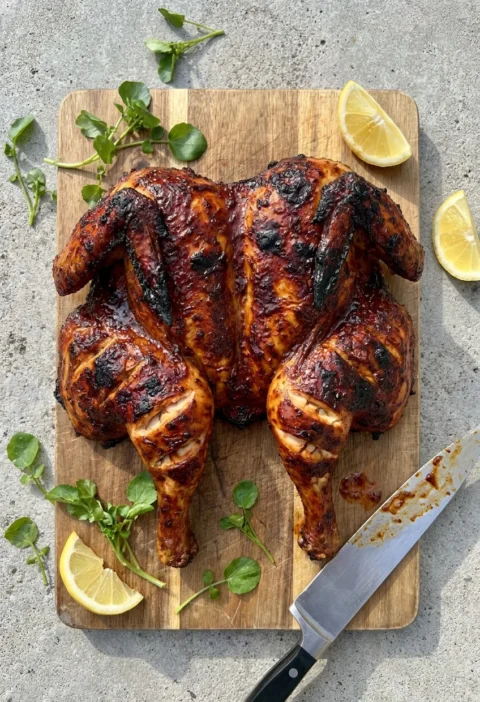 Spatchcocked piri piri chicken with dark charred marinade on a wooden chopping board with lemon wedges and watercress