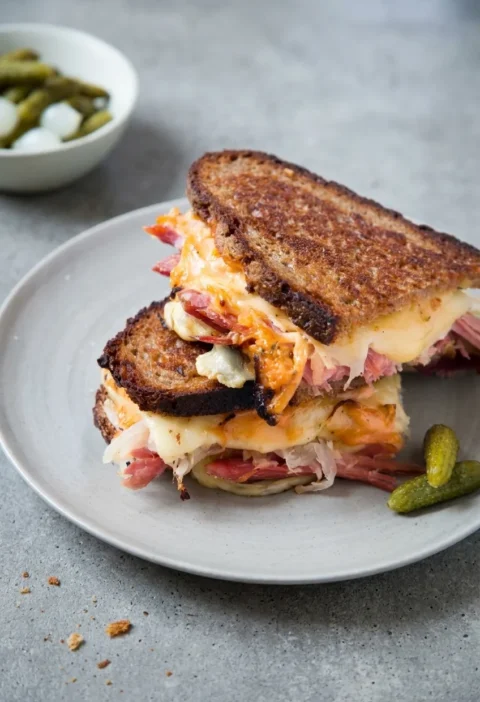 Hairy Bikers reuben sandwich cut in half and stacked, golden fried rye with melted Swiss cheese, salt beef and sauerkraut, cornichons on the plate