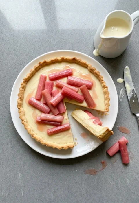 Hairy Bikers rhubarb and custard tart with pink forced rhubarb on set vanilla custard, one slice cut showing the layers