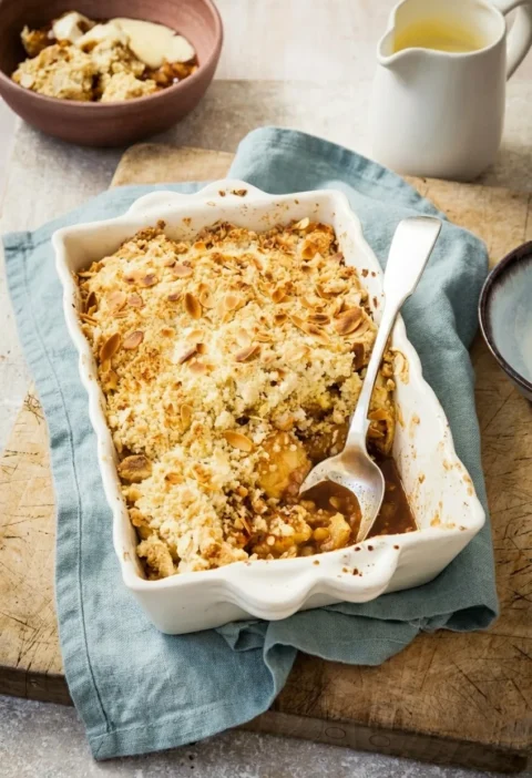 Hairy Bikers apple crumble in a fluted white dish with flaked almonds toasted on the golden topping and apple pieces showing where a spoon has been