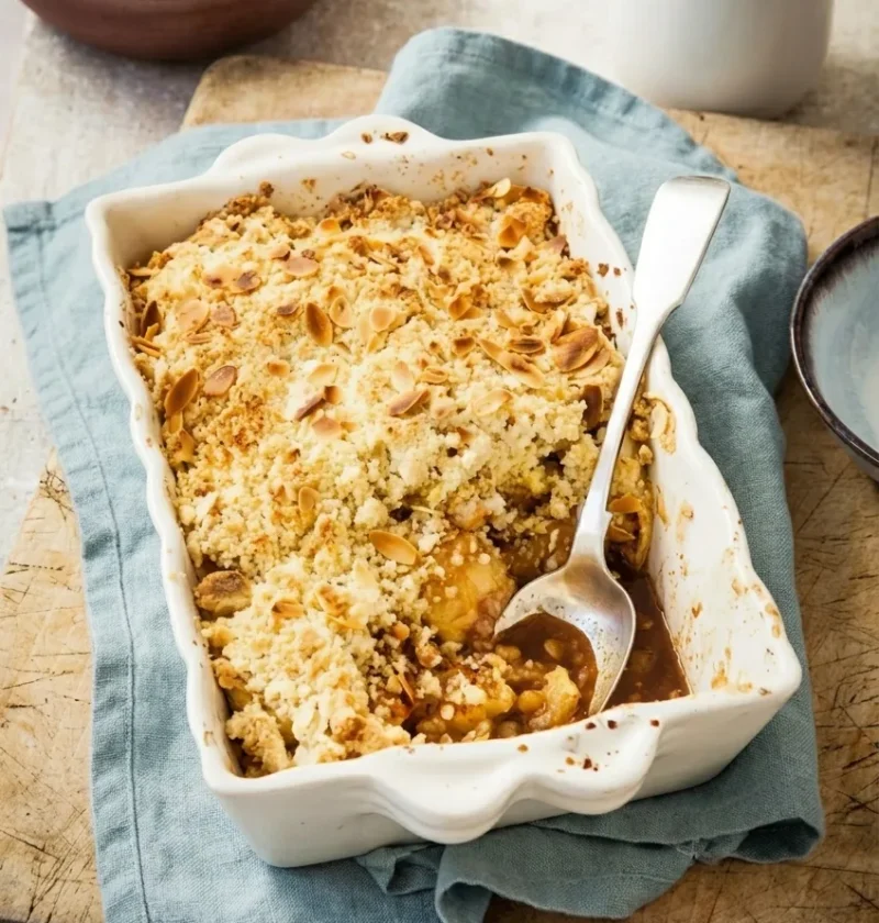 Hairy Bikers apple crumble in a fluted white dish with flaked almonds toasted on the golden topping and apple pieces showing where a spoon has been