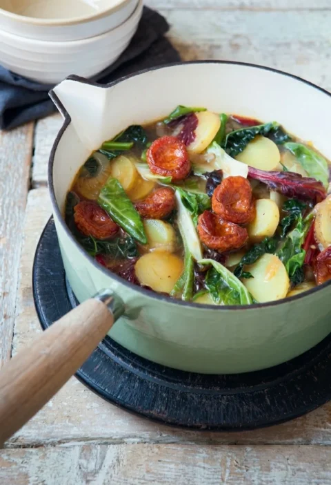Hairy Bikers caldo verde in a casserole pan with chorizo slices, halved waxy potatoes and shredded greens in a golden broth