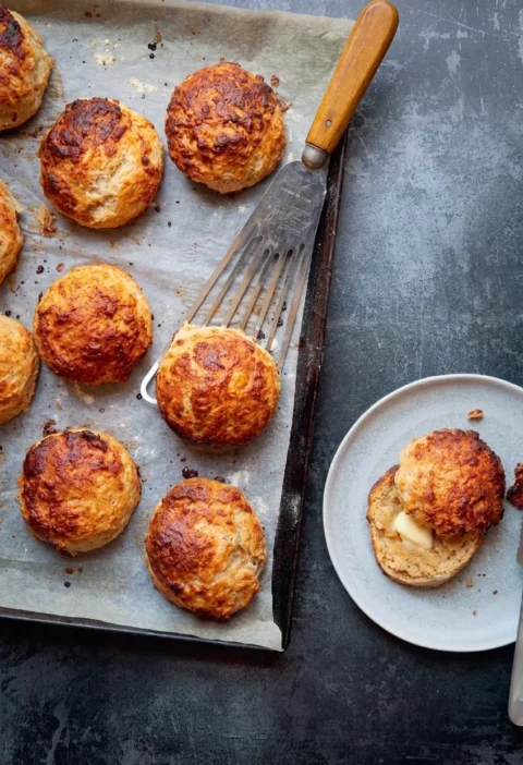 Hairy Bikers cheese and Marmite scones on a lined baking tray, deep golden and craggy, with one split and buttered on a plate
