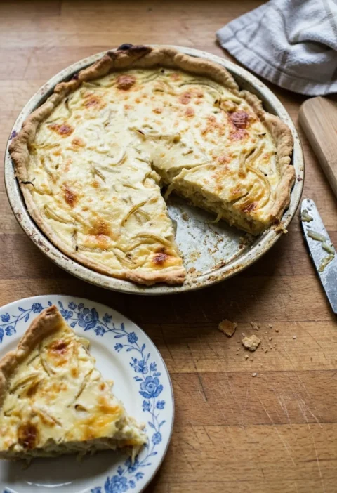 Hairy Bikers cheese and onion quiche in a flan tin with a wedge cut out, golden onion strands through a creamy custard under a patchy cheese top