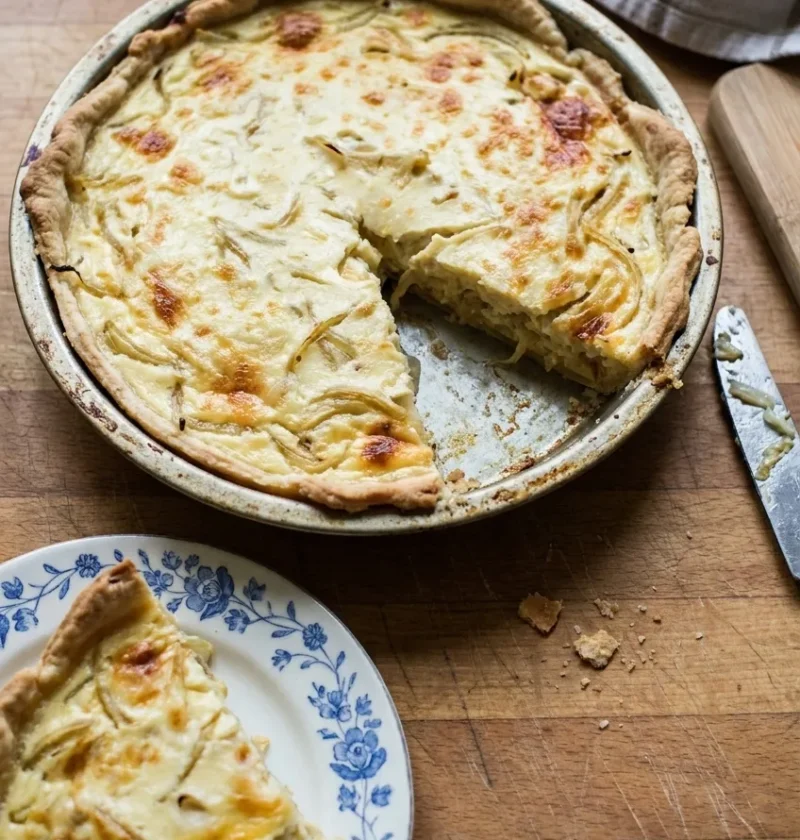 Hairy Bikers cheese and onion quiche in a flan tin with a wedge cut out, golden onion strands through a creamy custard under a patchy cheese top