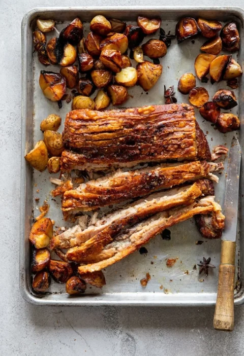 Hairy Bikers Chinese pork belly sliced on a baking tray, with crisp crackling, roast potatoes, whole garlic cloves and star anise