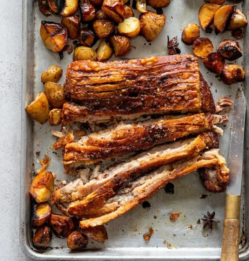 Hairy Bikers Chinese pork belly sliced on a baking tray, with crisp crackling, roast potatoes, whole garlic cloves and star anise