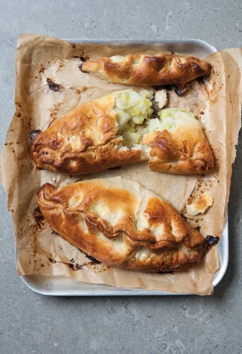 Hairy Bikers Cornish pasty with golden crimped pastry, one broken open on a baking tray to show the potato and swede filling