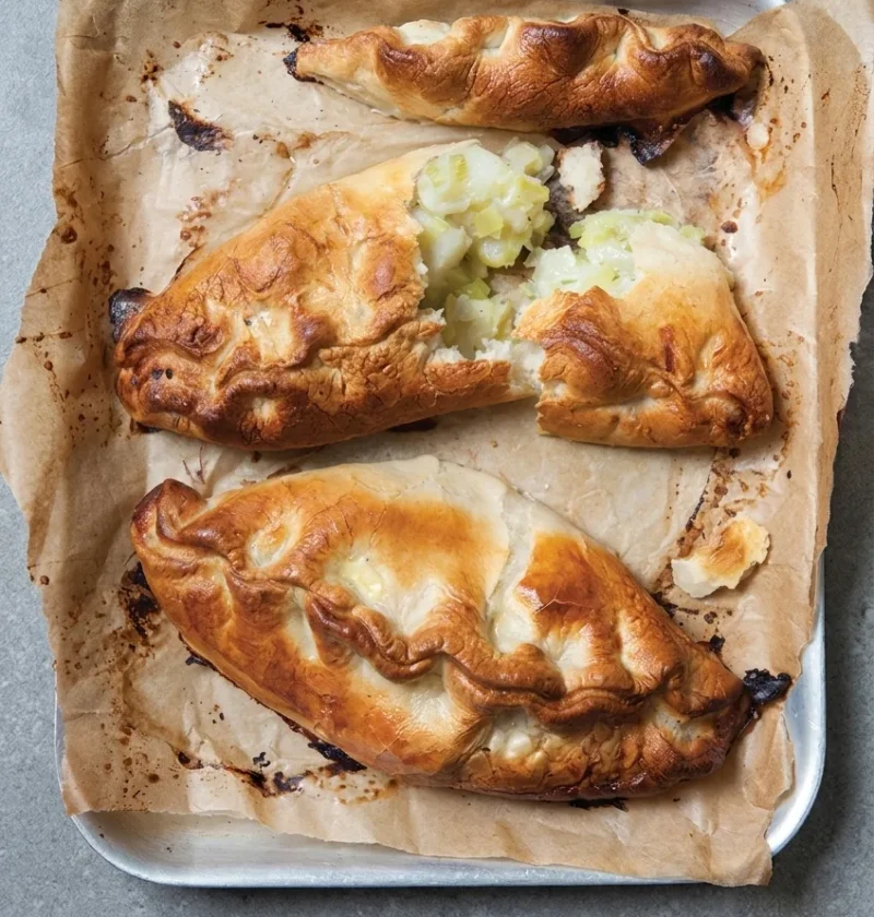 Hairy Bikers Cornish pasty with golden crimped pastry, one broken open on a baking tray to show the potato and swede filling