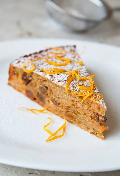 Hairy Bikers diet carrot cake, a slice showing grated carrot and sultanas through the crumb, dusted with icing sugar and topped with pared orange zest
