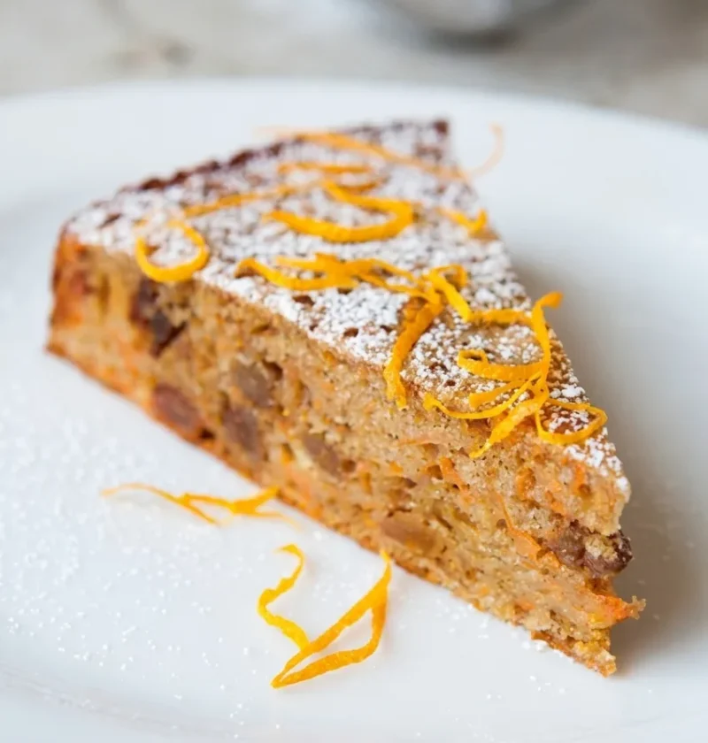 Hairy Bikers diet carrot cake, a slice showing grated carrot and sultanas through the crumb, dusted with icing sugar and topped with pared orange zest
