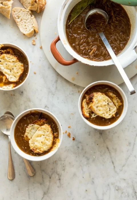 Hairy Bikers french onion soup in white bowls topped with Gruyere croutons, with a pot of caramelised onion soup alongside