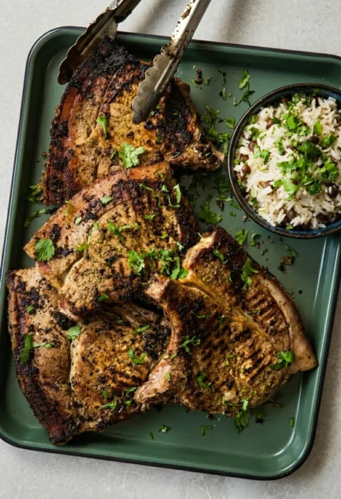 Hairy Bikers jerk pork chops on a green tray, four bone-in chops charred at the edges under a jerk marinade, with rice and peas alongside