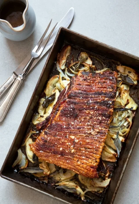 Hairy Bikers pork belly with crisp scored crackling, roasting on a bed of sliced apples, onions and sage