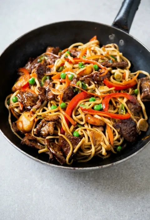 Hairy Bikers pork chow mein in a wok, with egg noodles, seared pork strips, red pepper, carrot, peas and water chestnuts in a glossy soy sauce