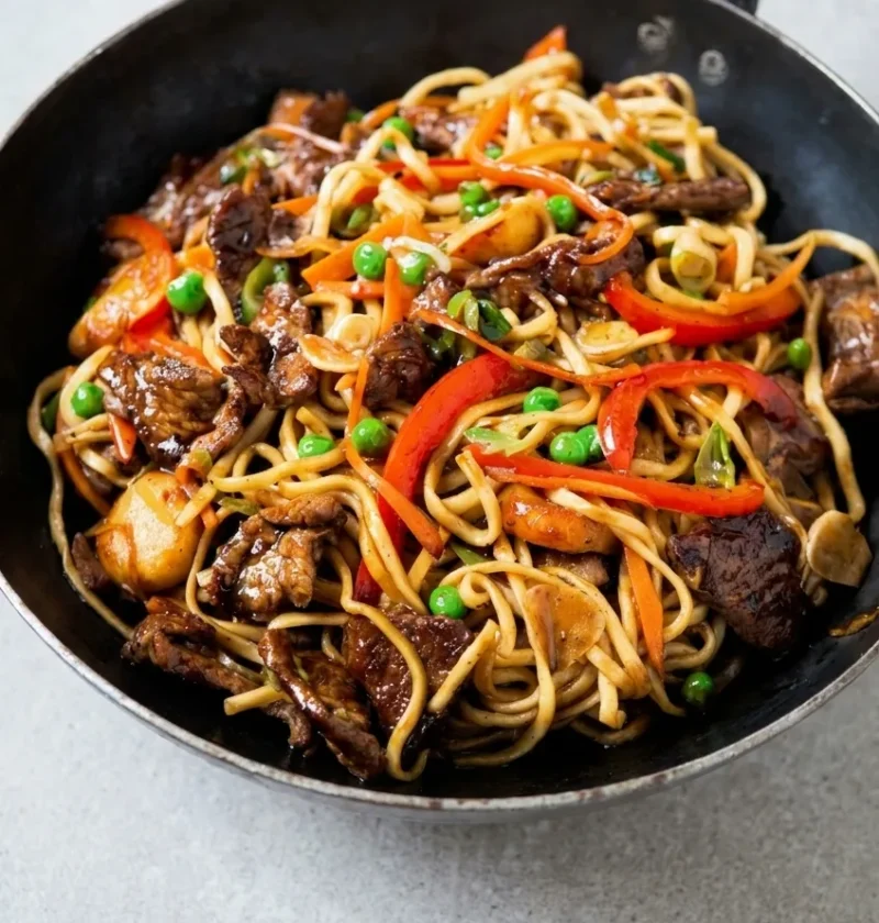 Hairy Bikers pork chow mein in a wok, with egg noodles, seared pork strips, red pepper, carrot, peas and water chestnuts in a glossy soy sauce