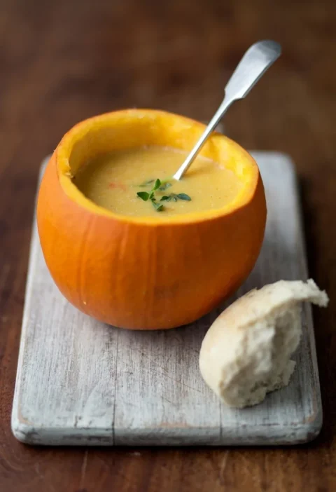 Hairy Bikers pumpkin soup served inside a hollowed-out pumpkin on a pale wooden board with a bread roll