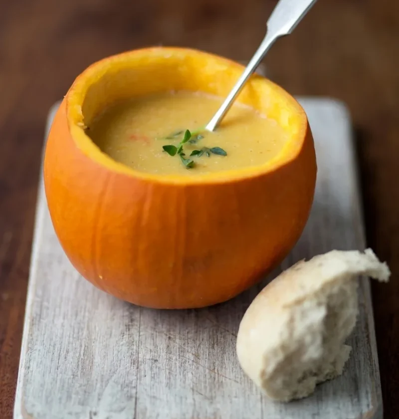 Hairy Bikers pumpkin soup served inside a hollowed-out pumpkin on a pale wooden board with a bread roll
