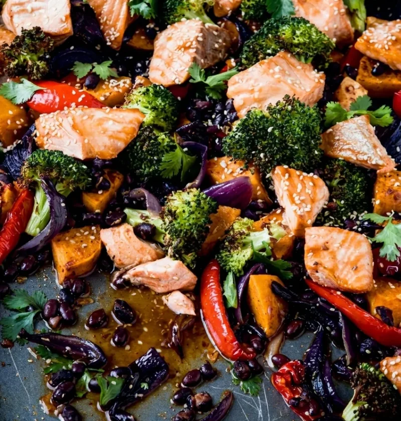 Hairy Bikers salmon tray bake in a roasting tin with pale pink salmon chunks on charred broccoli, sweet potato, red pepper and black beans
