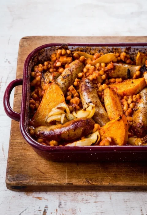 Hairy Bikers sausage and bean casserole in a burgundy roasting tin with browned sausages, butternut squash wedges, onion and haricot beans