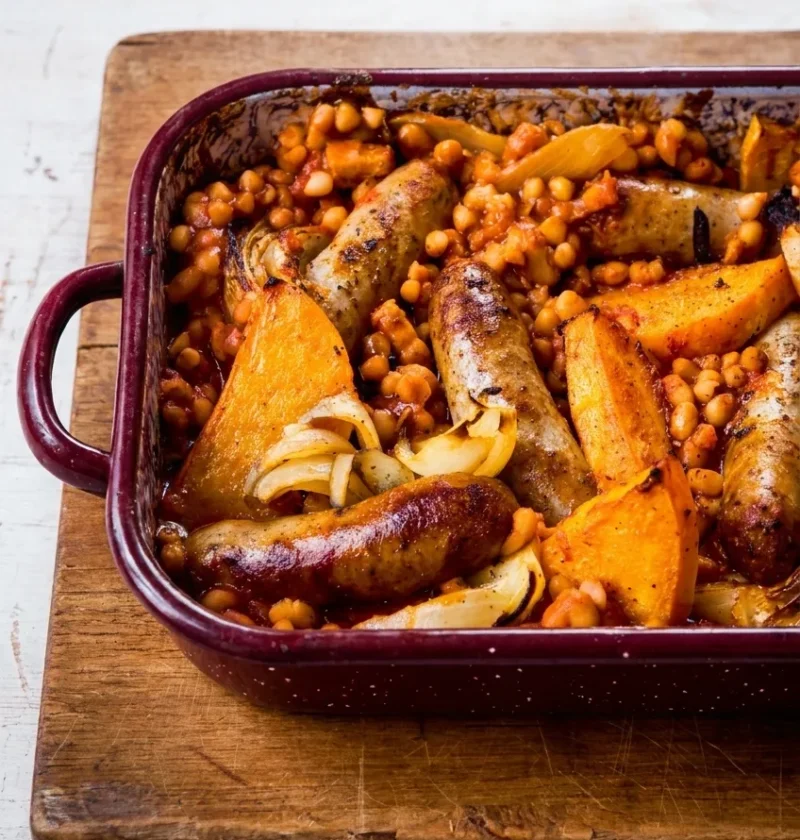 Hairy Bikers sausage and bean casserole in a burgundy roasting tin with browned sausages, butternut squash wedges, onion and haricot beans