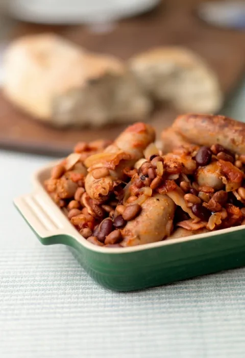 Hairy Bikers sausage casserole in a green dish with whole browned sausages, bacon, onion and beans in a thick tomato sauce