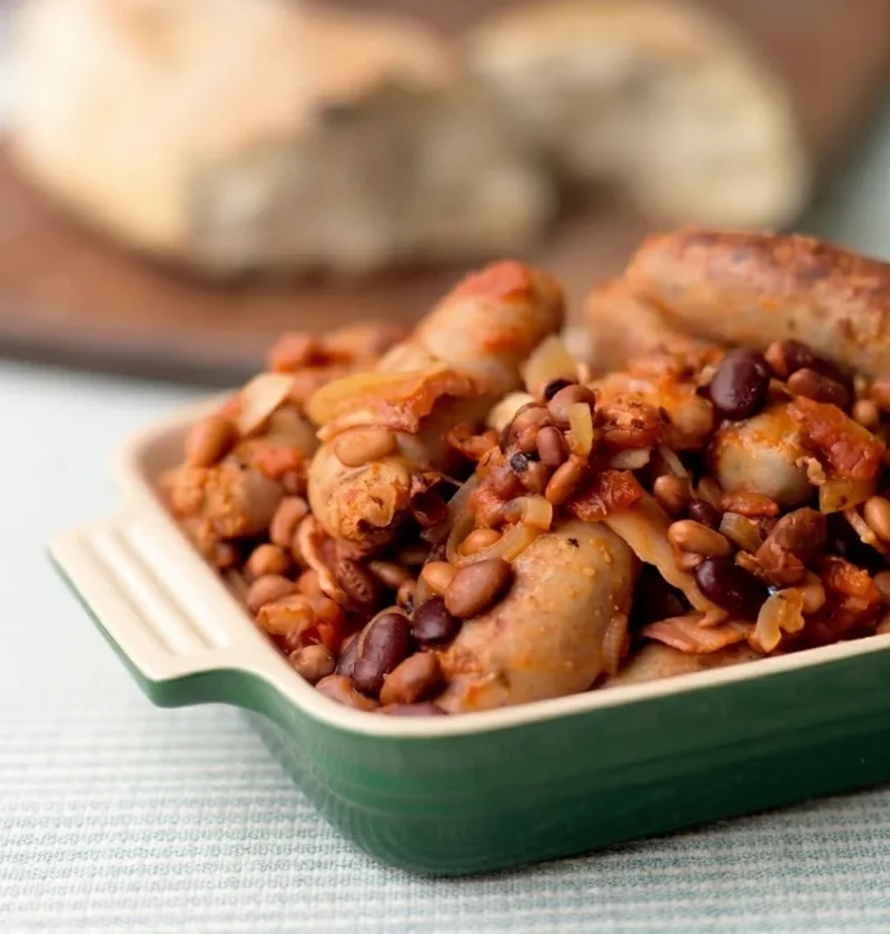 Hairy Bikers sausage casserole in a green dish with whole browned sausages, bacon, onion and beans in a thick tomato sauce