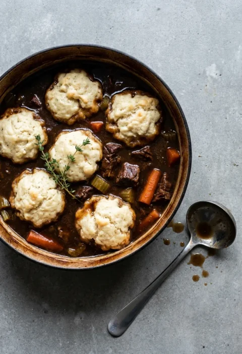 Hairy Bikers beef stew and dumplings in a cast iron pot, six risen suet dumplings sitting on dark Guinness gravy with chunks of beef and carrot