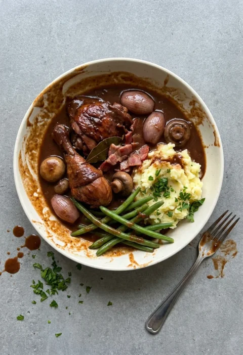 Hairy Bikers coq au vin in a white bowl, chicken and whole shallots in dark red wine sauce with mash