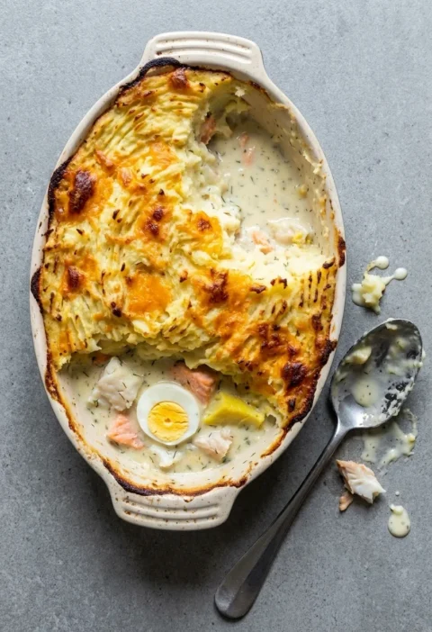Fish pie in an oval dish with browned cheesy mash, one portion scooped out showing salmon, white fish and a halved egg in dill sauce