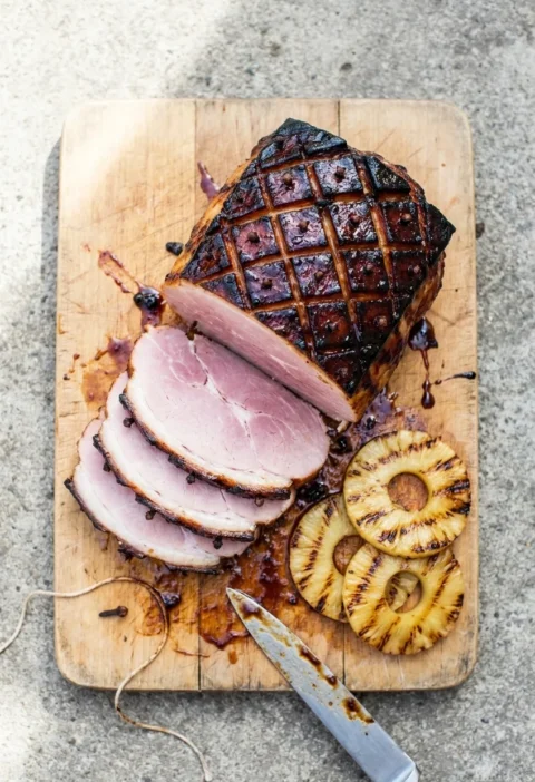 Glazed gammon joint with dark tamarind glaze and cloves, sliced on a wooden board with grilled pineapple rings