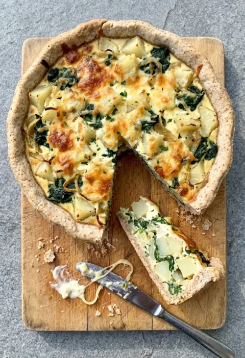 Open homity pie in a wholemeal crust with potato, spinach and browned Cheddar on a wooden board, one wedge cut out