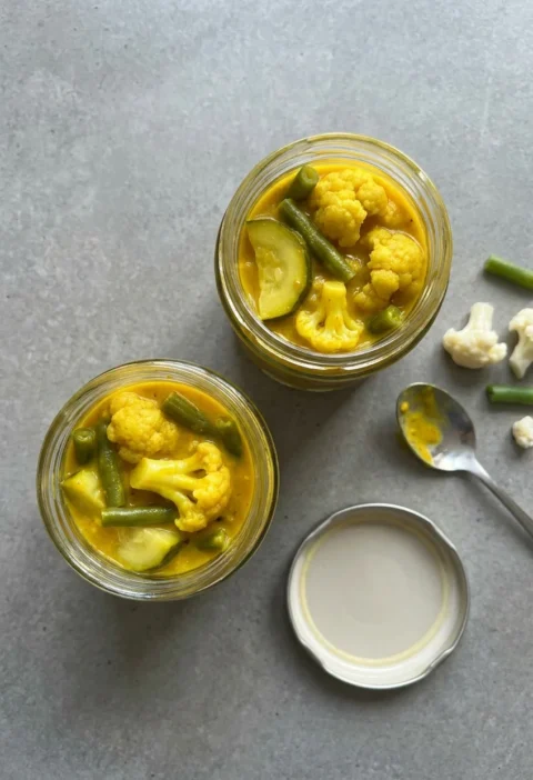 Hairy Bikers piccalilli in two open glass jars, thick bright yellow mustard pickle packed with cauliflower florets, green beans and courgette