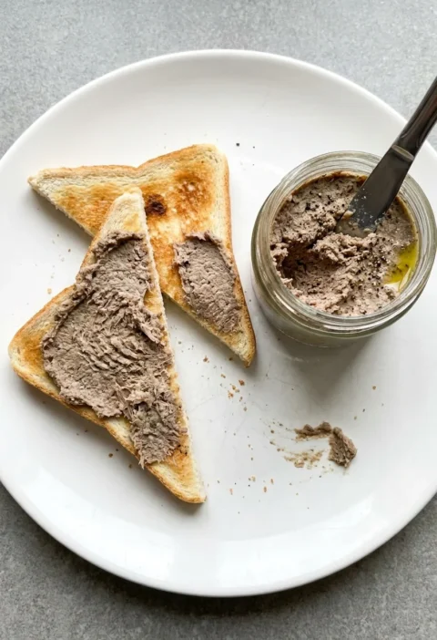 Coarse grey-brown potted beef spread thickly on triangles of white toast on a white plate, with a jar of the paste and a knife stuck in it