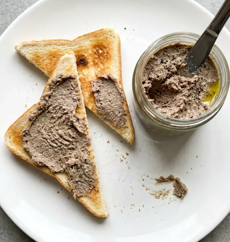 Coarse grey-brown potted beef spread thickly on triangles of white toast on a white plate, with a jar of the paste and a knife stuck in it