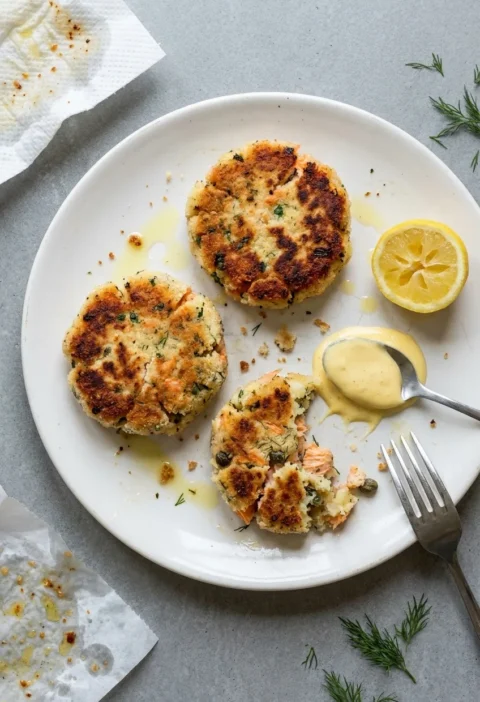 Salmon fish cakes with a patchy golden breadcrumb crust on a white plate, one broken open to show pink salmon flakes and dill through the mash, with hollandaise and a lemon wedge